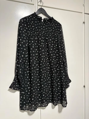 Svart blommig kortklänning med volang - Söt svart kortklänning med små vita blommor och volangdetaljer vid ärmslut och hals. Klänningen har lång ärm och lätt rynkad överdel för en avslappnad passform. Perfekt för dig som gillar en romantisk och trendig stil.