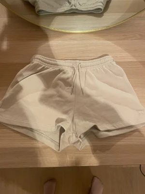 Beige sweatshorts med snörning - Säljer ett par beige sweatshorts med elastisk midja och snörning i storlek 146-152 . Shortsen har en avslappnad passform och är tillverkade i mjuk bomull. Liten broderad logga på benet för en clean look. Perfekta för sommardagar eller chill hemma.skriv för fler bilder, pris kan diskuteras!💗