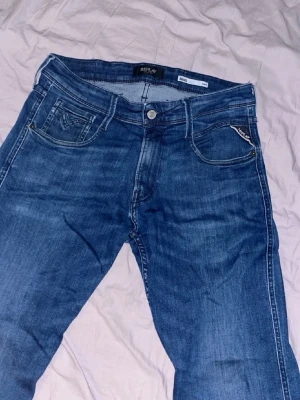 Blå jeans från Replay, slim fit - Snygga blå jeans från Replay i modellen Anbass med slim fit. Klassisk femficksdesign, diskreta slitningar och logga på myntfickan. Perfekta för dig som gillar en modern och stilren look.