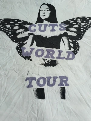Vit Guts World Tour t-shirt - Säljer en vit t-shirt med tryck av en person med fjärilsvingar och texten 'Guts World Tour' i lila. T-shirten har en avslappnad passform och korta ärmar. Perfekt för dig som gillar coola bandtröjor och statement-plagg.