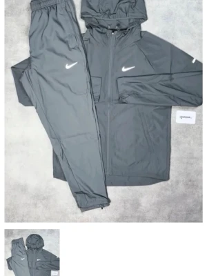 Grått Nike träningsset med huva - Snyggt grått träningsset från Nike med matchande jacka och byxor. Jackan har dragkedja, huva och vita Nike-loggor på bröstet och byxbenet. Tillverkat i lätt och funktionellt material som passar perfekt till träning eller sportaktiviteter.