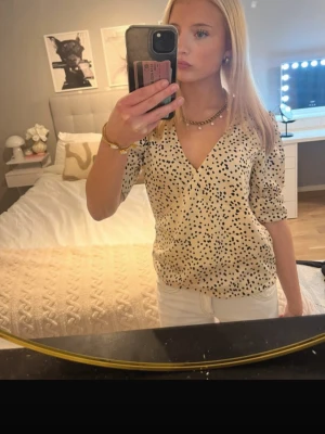 Beige prickig omlottblus - Trendig beige omlottblus med svarta prickar och v-ringning. Blusen har trekvartsärm och en avslappnad passform som ger en snygg siluett. Perfekt att matcha med jeans eller kjol för en cool look.