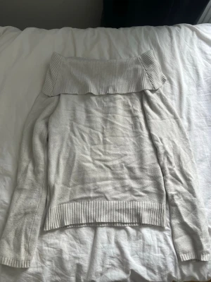 Ljusgrå off shoulder stickad tröja - Trendig ljusgrå stickad tröja med bred off shoulder-krage och långa ärmar. Tröjan har ribbstickade muddar vid ärmslut och nederkant samt en bred ribbad kant upptill som ger en snygg detalj. Perfekt för en avslappnad men stilren look.