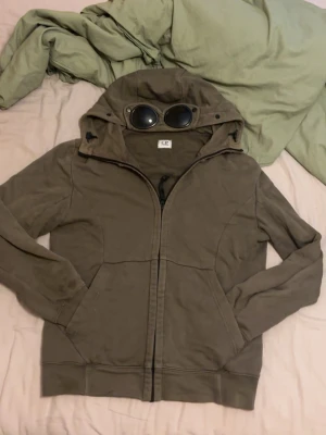Olivgrön hoodie med glasögon i huvan - Säljer en olivgrön hoodie från C.P. Company med dragkedja framtill och ikoniska glasögon i huvan. Hoodien har ribbade muddar, två sidofickor och justerbar dragsko nedtill. Perfekt för dig som gillar streetwear och unika detaljer.