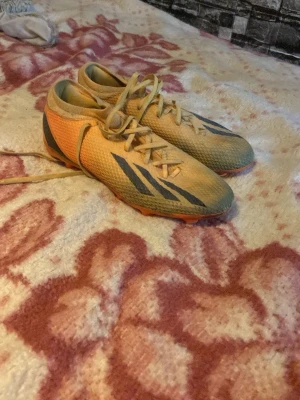 Gula fotbollsskor med svarta detaljer - Säljer ett par gula fotbollsskor med svarta detaljer och orange sula. Skorna har snörning och är tillverkade i ett lätt syntetmaterial för bra bollkontroll. Perfekta för dig som vill sticka ut på planen med snygg design och bra grepp.