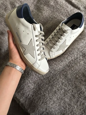 Vita sneakers med stjärndetalj - Snygga vita sneakers från Golden Goose med grå mockadetaljer och ikonisk stjärna på sidan. Skorna har mörkblå hälkappa, vita snören och beige sula. Tillverkade i läder och mocka för en cool och avslappnad look.