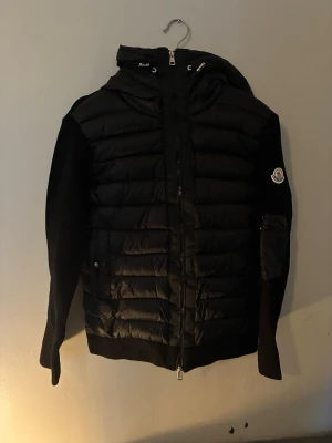Svart pufferjacka från Moncler - Snygg svart pufferjacka från Moncler med quiltad front och huva. Jackan har stickade ärmar och Moncler-logga på vänster ärm. Dragkedja framtill och fickor med knappstängning. Perfekt för kalla vinterdagar och riktigt trendig look. L - men egentligen M-S. Säljer för att den är för liten🔥 - vid snabba köp kan priset sänkas🔥