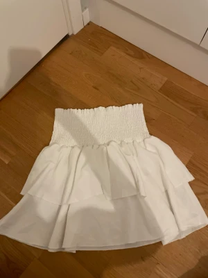 Vit volangkjol med smock - Supersöt vit kortkjol med smock i midjan och dubbla volanger. Kjolen har en luftig och lätt känsla, perfekt för varma dagar. Passar till både sneakers och sandaler. Enkel att matcha med olika toppar för en trendig look.