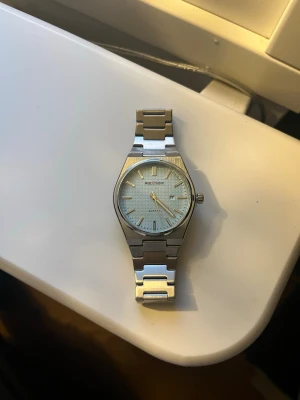 Silverfärgad klocka - Stilren klocka med silverfärgat armband i metall och ljusblå urtavla med diskret rutmönster. Klockan har klassisk design med datumvisning och tydliga markeringar. Perfekt för dig som gillar en clean och modern look.
