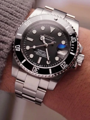 Seiko Submariner Mod ‘Black Steel’ 40 - -Movement: NH35 Seiko -Case Size: 40mm -Crown: Screw-down -Water Resistance: 300m -Crystal: Sapphire Glass -Lume: Super Lume -Bracelet: Full link bracelet included  Snygg Seiko Submariner-inspirerad klocka med svart urtavla, datumfönster och tydliga vita markeringar. Boetten och länken är i silverfärgat metall, vilket ger en klassisk och tidlös look. Perfekt för dig som gillar stilrena accessoarer.