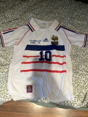 Frankrike VM 98 tröja Adidas M - Säljer en ikonisk vit fotbollströja från Adidas, officiell Frankrike VM 98 med nummer 10. Tröjan har blå och röda detaljer, krage och broderade loggor. Tillverkad i lätt och ventilerande Climalite-material. Perfekt för dig som älskar retro fotbollsstil.