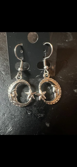 Silvriga örhängen med måne och stjärna - Unika silvriga örhängen i metall med hängande motiv av en måne och en stjärna. Månarna har ansikten och detaljerade kanter, vilket ger en cool och mystisk vibe. Perfekta för dig som gillar statement-smycken och vill sticka ut.