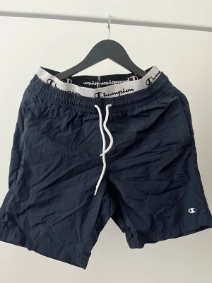 Marinblå shorts från Champion - Snygga marinblå shorts från Champion med vit snörning i midjan och elastiskt midjeband med logotyp. Perfekta för sommaren och har en sportig vibe. Lätta och bekväma att bära, passar till både chill och aktivitet.
