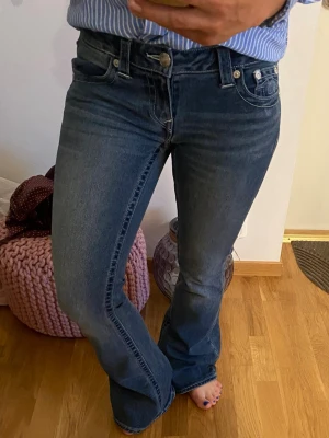 Blå bootcut jeans från True Religion - Säljer ett par blå bootcut jeans från True Religion med klassiska kontrastsömmar och broderade bakfickor. Jeansen har låg midja, fem fickor och är tillverkade i ett mjukt denimtyg med lätt stretch. Perfekta för en avslappnad men snygg look.