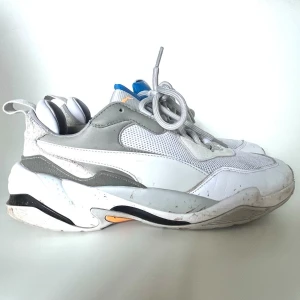 Puma Thunder Spectra - Chunky sneakers från Puma i vitt med grå och svarta detaljer samt inslag av orange och blått. Ovandelen är i mesh och syntetläder, med tjock sula och sportig design. Perfekta för dig som gillar streetwear och en trendig look.
