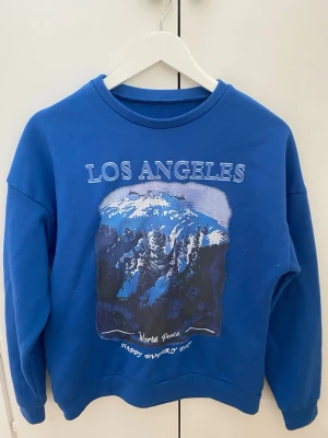  Los Angeles hoodie  - Blå hoodie med stort tryck av berg och texten 'Los Angeles' på bröstet. Mjuk insida och avslappnad passform, perfekt för en chill look. Rund halsringning och långa ärmar. Trycket har vita och mörkblå detaljer.