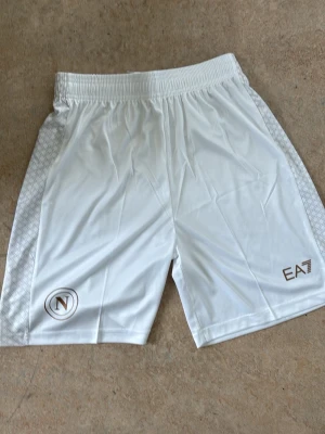 Vita EA7 shorts med logga - Snygga vita shorts från EA7 med elastisk midja och diskret mönstrade sidor. Shortsen har två loggor framtill, en EA7-logga och en rund logga med N. Perfekta för sport eller chill. Tillverkade i ett lätt och svalt syntetmaterial.