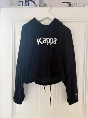 Svart hoodie från Kappa - Säljer en svart hoodie från Kappa med stor vit logga på bröstet och broderad logga på ärmen. Hoodien har dragsko i nederkanten och är gjord i mjukt, sammetsliknande material.