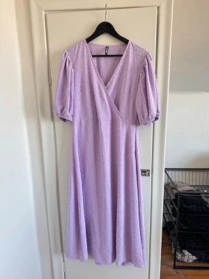 Ljuslila omlottklänning från Pieces - Säljer en ljuslila långklänning från Pieces med omlottmodell och v-ringning. Klänningen har puffiga korta ärmar med resår och ett diskret prickigt strukturmönster i tyget. Perfekt för sommarens alla tillfällen!