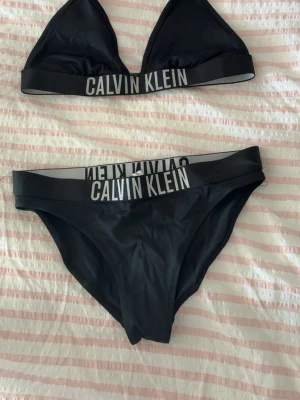 Svart Calvin Klein bikini - Snyggt svart bikini set från Calvin Klein med sportig känsla. Setet består av en trekants-bh och matchande trosor, båda med bred resår och tydlig logga i vitt. Materialet är stretchigt och bekvämt, perfekt för en modern och avslappnad stil. Det är storlek 14-16 men är väll i preferensen en xs