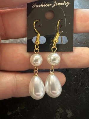 Guldörhängen med vita pärlor - Säljer ett par eleganta örhängen med guldfärgade krokar och dubbla vita pärlor, en rund och en droppformad. Perfekta för dig som vill ha en klassisk men ändå trendig accessoar. Materialet är metall och konstpärlor.