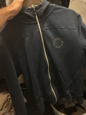 Mörkblå zip-hoodie från BOSS - Mörkblå hoodie med dragkedja från BOSS. Klassisk design med huva och vit logotyp på bröstet. Hoodien har en stilren look och är perfekt för en avslappnad stil. Materialet känns mjukt och bekvämt.
