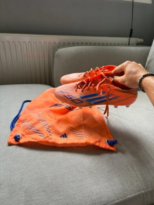 Adidas F50 FG – Fotbollsskor (inkl. originalpåse) - Säljer ett par Adidas F50 FG i den snygga orange/blå färgkombinationen. Skorna är i storlek 42 2/3 och är i fint begagnat skick med normalt slitage från användning, men har gott om liv kvar. Originalpåsen medföljer.  Perfekta för dig som vill ha en lätt och snabb fotbollssko med bra känsla och grepp på naturgräs eller konstgräs (FG).  Hör gärna av dig vid frågor eller om du vill ha fler bilder!