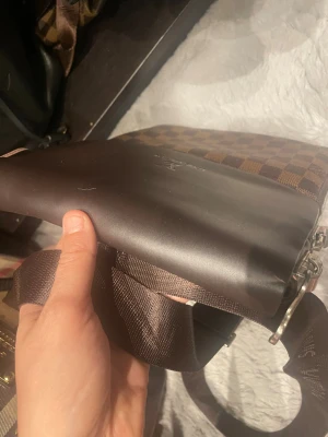 Brun axelväska med rutigt mönster - Snygg brun axelväska i konstläder med klassiskt rutigt mönster och bred axelrem. Väskan har ett lock och dragkedja upptill samt detaljer i silverfärg. Perfekt för dig som gillar stilrena accessoarer med en twist.