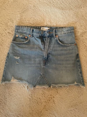 Blå jeanskjol från Zara - Säljer en blå jeanskjol från Zara med rå, fransig kant och slitna detaljer. Kjolen har klassisk femficksmodell, bälteshällor och dragkedja fram. Perfekt för dig som gillar en avslappnad och trendig stil.