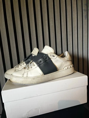 Valentino Garavani vita sneakers - Snygga vita sneakers från Valentino Garavani med bred svart rem över vristen och nitar på hälen. Skorna är i läder och har en chunky sula. Perfekta för dig som gillar en clean men ändå edgy stil.