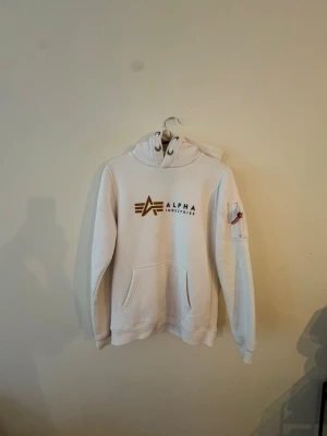 Vit hoodie Alpha Industries - Snygg vit hoodie från Alpha Industries med guldigt logotryck på bröstet och ficka på ärmen med röd detalj. Klassisk känguruficka framtill och huva med dragsko. Perfekt för en avslappnad och trendig stil.