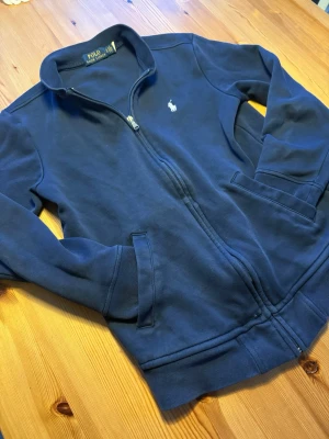 Mörkblå zip hoodie från Polo Ralph Lauren - Snygg mörkblå hoodie med dragkedja från Polo Ralph Lauren. Klassisk design med två fickor framtill och den ikoniska broderade loggan på bröstet. Tillverkad i mjukt bomullsmaterial och har ribbade muddar vid ärmslut och nederkant. Perfekt för en avslappnad stil.