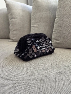 Svart kuvertväska med paljetter - Snygg svart kuvertväska täckt med glansiga paljetter. Väskan har jag virkat själv och har därför en unik form som gör den till ett statement piece. Perfekt för dig som vill sticka ut och addera lite extra bling till din outfit.
