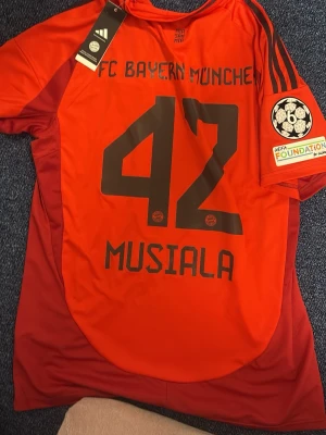 Bayern München Musiala matchtröja - Snygg röd Bayern München matchtröja från Adidas med Musiala och nummer 42 på ryggen. Tröjan har svarta detaljer, klubbmärke och Adidas-logga på bröstet samt Champions League-märke på ärmen. Tillverkad i lätt och ventilerande material.