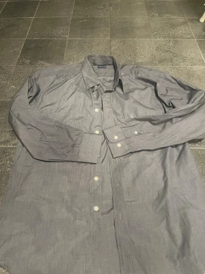 Grå skjorta från Ralph Lauren - Snygg grå skjorta från Ralph Lauren med klassisk krage och vita knappar. Skjortan har lång ärm och diskret broderad logga på bröstet. Tillverkad i bomull för en bekväm känsla och stilren look. Storlek är L/M