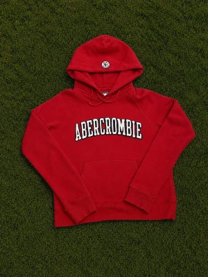 Röd hoodie från Abercrombie & Fitch - Det står M men passar Xs.                      Snygg röd hoodie från Abercrombie & Fitch med stor vit logga på bröstet. Klassisk känguruficka framtill och huva med dragsko. Tillverkad i mjuk bomullsblandning (72% bomull, 28% polyester) för skön känsla. Perfekt för en avslappnad och trendig look.