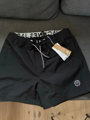Svarta badbyxor Jack & Jones  - Snygga svarta badbyxor från Jack & Jones med vit dragsko i midjan och elastisk linning. Diskret logga med palmtryck på ena benet och meshfoder på insidan för extra komfort. Perfekta för stranden eller poolen. Köpta för 300kr