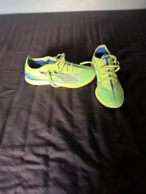 Neongröna futsalskor från Puma - Säljer ett par neongröna futsalskor från Puma med blå och vita detaljer. Skorna har en sportig design, snörning och slitstark sula med bra grepp. Perfekta för dig som vill bli bättre på futsal eller bara vill ha en futsal pass