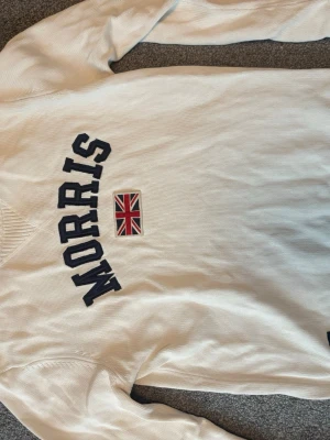 Vit tröja från Morris med tryck - Snygg vit tröja från Morris med mörkblå text och brittisk flagga broderad på bröstet. Tröjan har rund halsringning och ribbade muddar. Perfekt för dig som gillar klassisk och sportig stil.