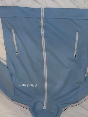 Blå ziptröja från Palm Angels - Snygg ljusblå ziptröja från Palm Angels med vita detaljer och dragkedja framtill. Tröjan har två fickor med dragkedja och vita ränder längs ärmarna. Perfekt för en sportig och avslappnad stil.