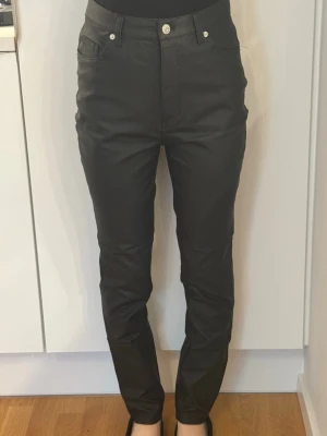 Svarta coated jeans från NA-KD - Snygga svarta coated jeans från NA-KD med hög midja och smal passform. Jeansen har klassisk femficksdesign, bälteshällor och en glansig finish som ger en cool look. Perfekta för dig som vill ha ett par trendiga och stilrena jeans. OBS! Modellen är XS och varan är i storlek S