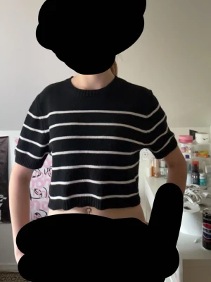 Svart stickad crop top med ränder - Säljer en svart stickad crop top med vita horisontella ränder. Tröjan har korta ärmar, rund halsringning och ribbade kanter. Perfekt för en avslappnad och trendig look. Materialet känns mjukt och stretchigt.