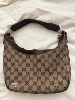 Gucci GG hobo bag - Säljer en klassisk beige axelremsväska från Gucci med det ikoniska GG-mönstret i brunt. Väskan har detaljer i mörkbrunt läder och en bred axelrem med grönt och rött band. Stängs med dragkedja och har silverfärgade metalldetaljer. Kom privat för närmare bilder