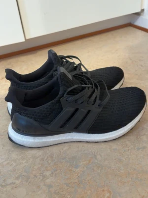 Adidas Ultra Boost svarta sneakers - Svarta Adidas Ultra Boost sneakers med stickad ovandel och vita Boost-sulor. Skorna har klassisk tre-randig design på sidorna, snörning och en bekväm, sportig look. Perfekta för dig som gillar streetstyle och sportig vibe.