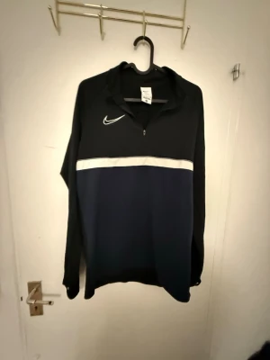 Nike Dri-FIT svart träningsjacka - Nike Dri-FIT träningsjacka i svart med vit rand över bröstet och dragkedja vid halsen. Tillverkad i funktionsmaterial som andas, perfekt för träning eller uppvärmning. Långärmad och stilren design med Nike-logga på bröstet.