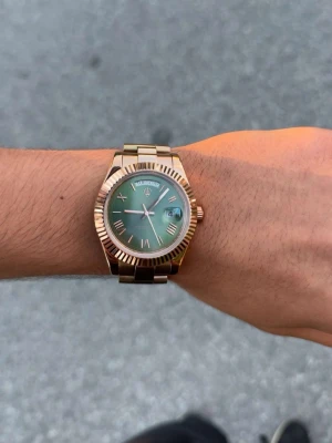 Guldig klocka med grön urtavla - Snygg klocka i guld med länkarmband och grön urtavla. Klockan har datumvisning och veckodag högst upp på urtavlan. Klassisk design med räfflad kant runt glaset och stilrena visare. Perfekt accessoar för att lyfta din outfit. Vatten tätt också