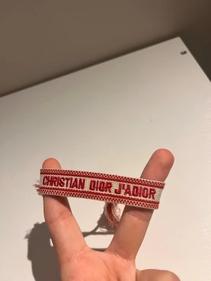 Christian Dior J'Adior armband röd/vit - Snyggt vävt armband från Christian Dior med texten 'J'Adior' i rött på vit botten. Armbandet har tofsar i ändarna och en cool, bohemisk känsla. Perfekt accessoar för att lyfta din outfit med en exklusiv touch.