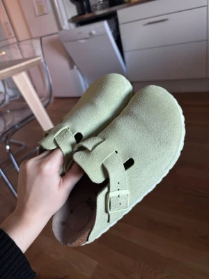 Ljusgröna mockatofflor Birkenstock - Säljer ett par ljusgröna tofflor från Birkenstock i mjuk mocka. Modellen har en bred rem med spänne och öppen häl, samt en bekväm fotbädd i kork. Perfekta för dig som vill ha både stil och komfort hemma eller ute.