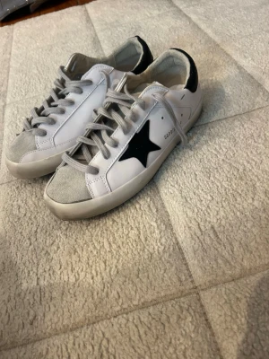 Golden goose skor  - Säljer ett par vita sneakers med svart stjärndetalj på sidan och grå mocka vid tån. Skorna har vita och grå partier, vita sulor och grå skosnören. Snygg och stilren design som passar till det mesta.