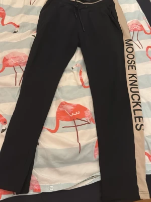 Svarta Moose Knuckles mjukisbyxor - Snygga svarta mjukisbyxor från Moose Knuckles med beige sidopanel och stor logga längs benet. Byxorna har dragsko i midjan och raka ben. Perfekta för en chill och avslappnad stil.